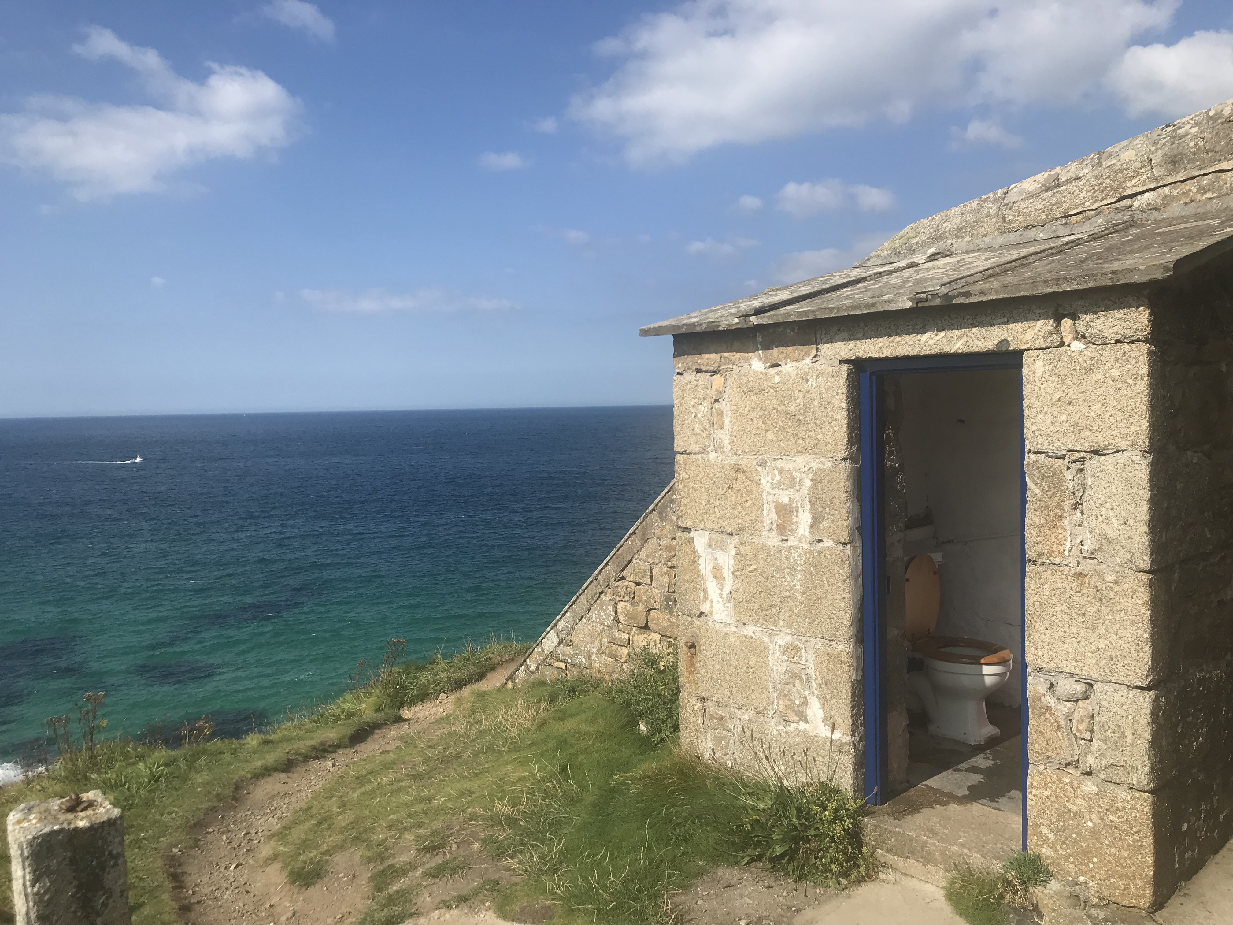 A toilet in a small stone building on a green hillside with blue sea and blue skies behind.